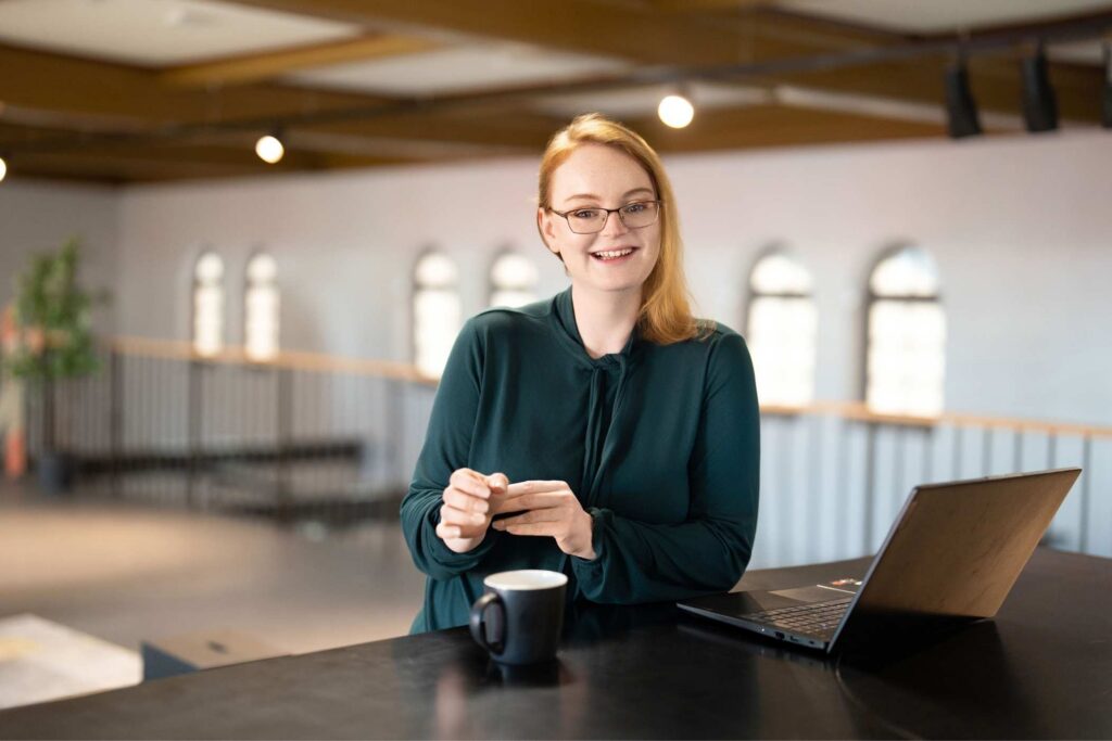 Julia, SEO-Partnerin für KMUs, steht freundlich lächelnd an einem Stehtisch mit Laptop und Kaffeetasse in einem hellen, modernen Büro.