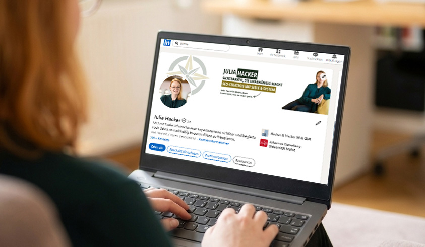 Eine Frau mit rötlichen Haaren sitzt von hinten im Anschnitt auf einem Sessel und tippt auf einem grauen Laptop. Der Bildschirm zeigt das LinkedIn Profil von Julia Hacker
