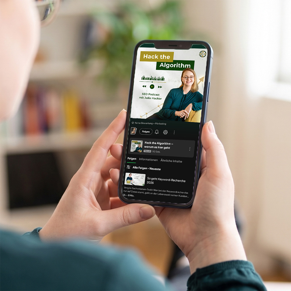 Eine Person hält ein Smartphone in beiden Händen. Auf dem Display ist eine Spotify geöffnet, die den Kanal "Hack the Algorithm – SEO Podcast mit Julia Hacker" anzeigt. Auf dem Bildschirm ist das Coverbild mit Julia Hacker in einer grünen Bluse sowie eine aktuelle Episode zum Thema Keyword-Recherche zu sehen.