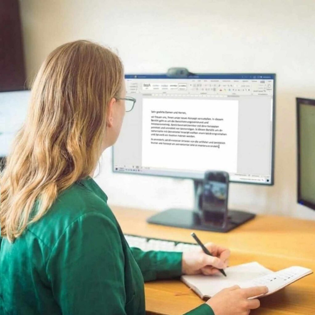 Julia Hacker Eine Rückansicht von Julia Hacker, die an einem Schreibtisch sitzt und in ein Notizbuch schreibt. Im Hintergrund befindet sich ein Desktop-Monitor, auf dem ein Textverarbeitungsdokument zu sehen ist.