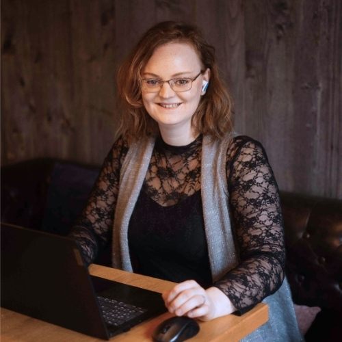 Julia Hacker sitzt an einem Holztisch in einem Raum mit rustikaler Holzwand und arbeitet an einem Laptop. Sie trägt ein schwarzes Oberteil mit Spitzenärmeln, eine graue Weste, eine Brille und einen In-Ear-Kopfhörer im rechten Ohr. Sie blickt auf den Bildschirm des Laptops, während ihre rechte Hand auf dem Gehäuse ruht. Vor ihr auf dem Tisch steht eine kabellose Maus, daneben eine kleine Topfpflanze. Im Hintergrund ist eine dunkle, gesteppte Lederbank zu sehen. Das Foto ist im Querformat und zeigt Julia Hacker von der Taille aufwärts.
