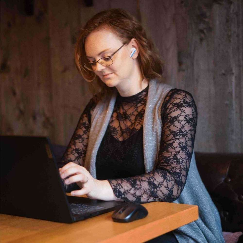Julia Hacker sitzt an einem Holztisch in einem Raum mit rustikaler Holzwand und arbeitet an einem Laptop. Sie trägt ein schwarzes Oberteil mit Spitzenärmeln, eine graue Weste, eine Brille und einen In-Ear-Kopfhörer im rechten Ohr. Sie blickt auf den Bildschirm des Laptops, während ihre rechte Hand auf dem Gehäuse ruht. Vor ihr auf dem Tisch steht eine kabellose Maus, daneben eine kleine Topfpflanze. Im Hintergrund ist eine dunkle, gesteppte Lederbank zu sehen. Das Foto ist im Querformat und zeigt Julia Hacker von der Taille aufwärts.