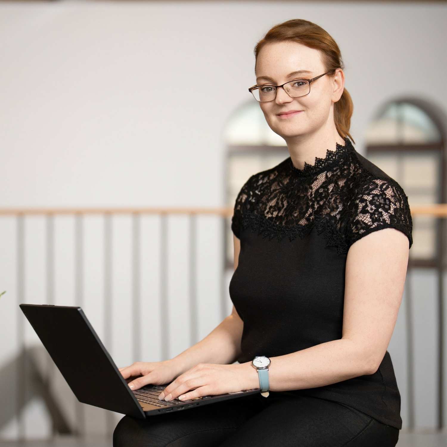 Ein professionelles Foto von Julia Hacker, Interim SEO Beraterin. Sie sitzt auf der rechten Seite des Bildes, blickt lächelnd in die Kamera und hält einen aufgeklappten Laptop auf ihrem Schoß. Sie trägt eine Brille und ein schwarzes Oberteil mit Spitzendetails. Die linke Bildhälfte ist ein heller, leerer Raum (Negativraum), und der Hintergrund zeigt ein modernes Gebäudeinterieur mit Fenstern.